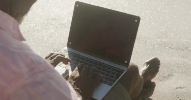 Senior african american man sitting and using laptop on sunny beach. healthy, active retirement beach holiday.
