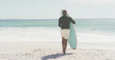 Kıdemli Afro-Amerikan adam güneşli sahilde sörf tahtasıyla yürüyor. sağlıklı, aktif emeklilik plaj tatili.