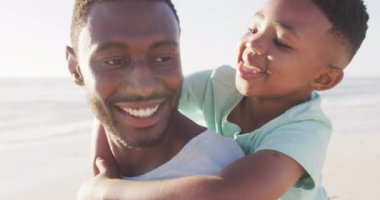 Gülümseyen Afro-Amerikan babası oğlunu güneşli sahilde taşıyor. sağlıklı, aktif aile plaj tatili.