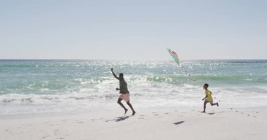 Gülümseyen Afrikalı Amerikalı baba ve oğlu güneşli sahilde uçurtma uçuruyorlar. sağlıklı, aktif aile plaj tatili.