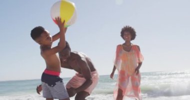 Gülümseyen Afro-Amerikan ailesi güneşli sahilde topla oynuyor. sağlıklı, aktif aile plaj tatili.