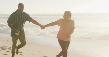 Gülümseyen kıdemli Afro-Amerikan çift el ele tutuşup güneşli sahilde koşuyor. sağlıklı, aktif emeklilik plaj tatili.