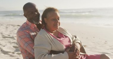 Gülümseyen son sınıf Afro-Amerikan çifti güneşli sahilde sarılıp oturuyorlar. sağlıklı, aktif emeklilik plaj tatili.