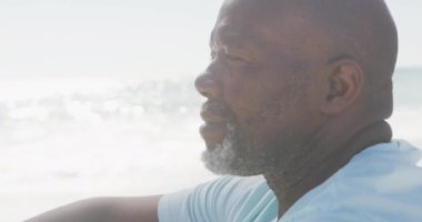 Beyaz t-shirt giyen son sınıf Afro-Amerikan erkeği güneşli sahilde oturuyor. sağlıklı, aktif emeklilik plaj tatili.