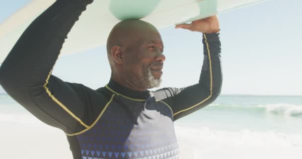 一个面带微笑的资深非洲裔美国人与冲浪板一起在阳光明媚的海滩上行走的画像 积极的退休海滩假期 — 图库视频影像
