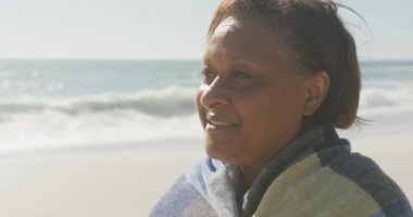 Güneşli sahilde battaniyeli kıdemli Afrikalı Amerikalı kadın gülümsüyor. sağlıklı, aktif emeklilik plaj tatili.