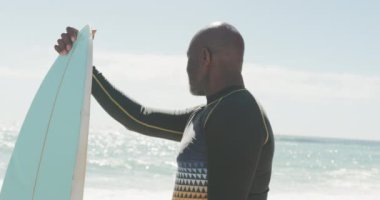 Kıdemli Afro-Amerikan adam güneşli sahilde sörf tahtasıyla ayakta duruyor. sağlıklı, aktif emeklilik plaj tatili.