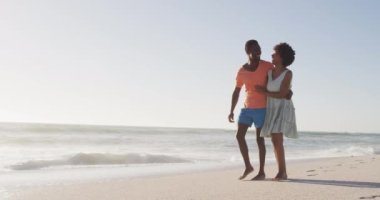 Smiling african american couple embracing and walking on sunny beach. healthy, active family beach holiday.