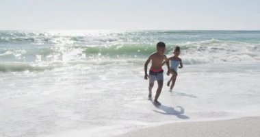 Smiling african american siblings running and wearing swimming suits on sunny beach. healthy, active family beach holiday.