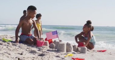 Gülümseyen Afro-Amerikan ailesi Sunny Beach 'te Amerikan bayrağıyla kumdan kale inşa ediyor. sağlıklı, aktif aile plaj tatili.