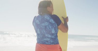 Kıdemli Afro-Amerikan kadın güneşli sahilde sörf tahtasıyla ayakta duruyor. sağlıklı, aktif emeklilik plaj tatili.