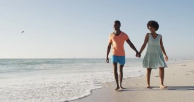 Gülümseyen Afro-Amerikan çifti el ele tutuşup güneşli sahilde yürüyor. sağlıklı, aktif aile plaj tatili.