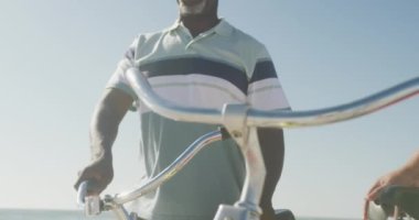 Smiling senior african american couple walking with bicycles on sunny beach. healthy, active retirement beach holiday.