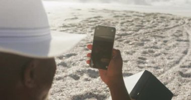 Kıdemli Afro-Amerikan adam güneşli sahilde güneşli yatakta uzanıyor ve akıllı telefon kullanıyor. sağlıklı, aktif emeklilik plaj tatili.