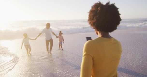 アフリカ系アメリカ人の家族が写真を撮り 日当たりの良いビーチを歩いて笑っています 健康的で活動的な家族のビーチホリデー — ストック動画