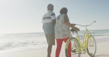 Kıdemli Afro-Amerikan çifti güneşli sahilde bisikletle yürüyor. sağlıklı, aktif emeklilik plaj tatili.