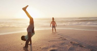 Smiling african american siblings raising hands and playing on sunny beach. healthy, active family beach holiday.
