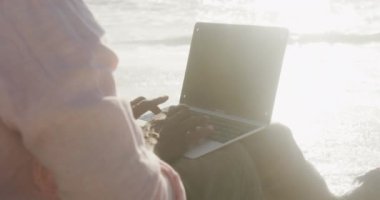 Senior african american man sitting and using laptop on sunny beach. healthy, active retirement beach holiday.