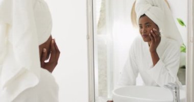 Video of happy african american woman in robe looking at mirror and moisturizing face. beauty treatment and skin care routine concept.