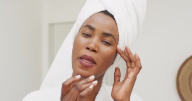 Video of happy african american woman in robe moisturizing face. beauty treatment and skin care routine concept.
