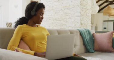 Video of happy african american woman on sofa with headphones using laptop. leisure and relax, spending free time with technology at home.