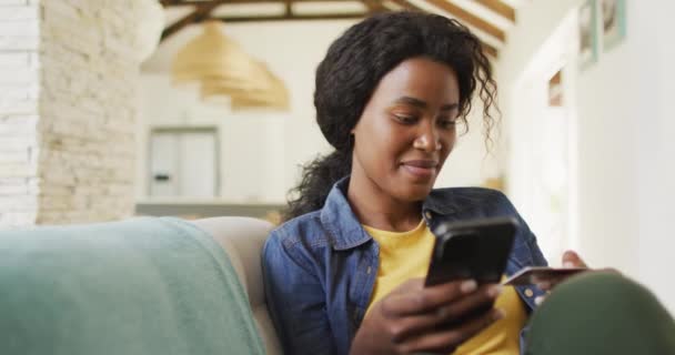 スマホを使ってソファで幸せなアフリカ系アメリカ人女性の動画 余暇とリラックスして家でテクノロジーと自由な時間を過ごし — ストック動画