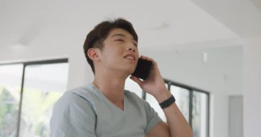 Asyalı genç erkek akıllı telefondan konuşuyor ve oturma odasında gülümsüyor. Evde yalnız zaman geçirmek..