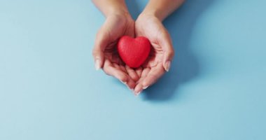 Video of hands of caucasian woman holding heart on blue background. medicine, health, cancer awareness concept digitally generated video.