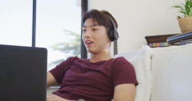 Mutlu Asyalı genç erkek kulaklık takıyor ve oturma odasında dizüstü bilgisayar kullanıyor. Evde yalnız zaman geçirmek..