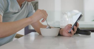 Asyalı genç erkek akıllı telefonuyla masada oturuyor ve kahvaltı yapıyor. Evde yalnız zaman geçirmek..