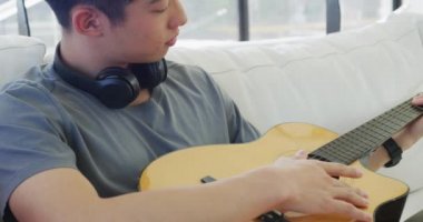 Kulaklık takan, gitar çalan ve oturma odasında oturan Asyalı bir genç. Evde yalnız zaman geçirmek..