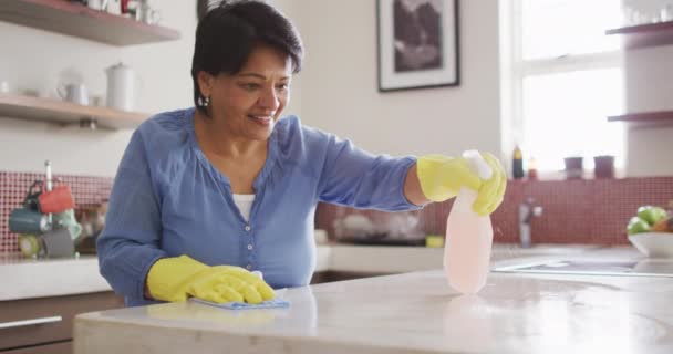 Smiling Senior Biracial Woman Wearing Gloves Cleaning Table Kitchen Alone — Wideo stockowe