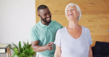 Kafkasyalı yaşlı kadını inceleyen mutlu Afrikalı Amerikalı erkek fizyoterapistin videosu. Yaşlılar sağlık ve huzurevi konsepti.