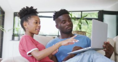 Kanepede oturup dizüstü bilgisayar kullanan mutlu Afro-Amerikan çiftin videosu. Aşk, ilişki, birliktelik ve evde teknolojiyle zaman geçirmek..