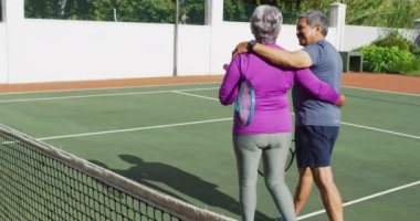 Çift ırklı mutlu çiftin tenis kortunda raketlerle sarılıp yürüdüğü bir video. aktif emeklilik yaşam tarzı, kıdemli ilişki ve tenis eğitim konsepti.
