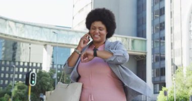 Mutlu ve çift ırklı bir kadın. Akıllı olanı kontrol ediyor ve şehirde yürüyor. dijital göçebe hareket halinde, beden içeriği.