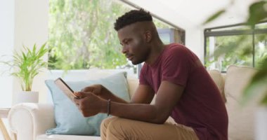 Video of relaxed african american man sitting on sofa and using tablet. relaxing and spending time with technology at home.