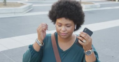 Mutlu ve çift ırklı kadın akıllı telefon kullanıyor ve şehirde kulaklık takıyor. dijital göçebe hareket halinde, beden içeriği.