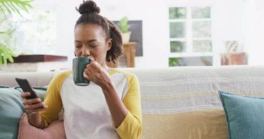 Afro-Amerikalı kadın kahve içiyor ve akıllı telefon kullanıyor. Evdeki kanepede oturuyor. Yaşam tarzı ve teknoloji kavramı