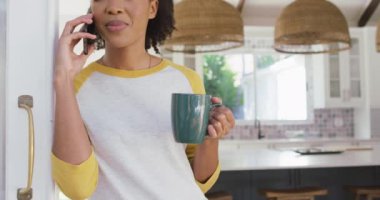 Afro-Amerikalı kadın elinde kahve fincanı tutarak evde akıllı telefondan konuşuyor. Yaşam tarzı ve teknoloji kavramı
