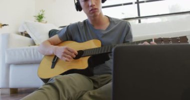 Kulaklık takan Asyalı çocuk evde dizüstü bilgisayara bakarak gitar çalıyor. Genç yaşam tarzı ve yaşam konsepti