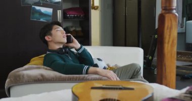 Asyalı çocuk akıllı telefondan konuşuyor. Evdeki kanepede oturuyor. Genç yaşam tarzı ve yaşam konsepti