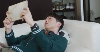 Asyalı çocuk evdeki kanepede uzanmış dijital tablette oyun oynuyor. Genç yaşam tarzı ve yaşam konsepti