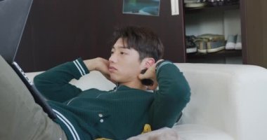 Kulaklık takan Asyalı çocuk evdeki kanepede dizüstü bilgisayar kullanıyor. Genç yaşam tarzı ve yaşam konsepti