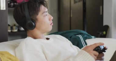 Kulaklık takan Asyalı çocuk evdeki kanepede oturmuş video oyunları oynuyor. Genç yaşam tarzı ve yaşam konsepti