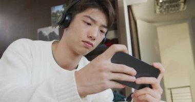 Kulaklık takan Asyalı çocuk akıllı telefonuyla oyun oynuyor evdeki kanepede oturuyor. Genç yaşam tarzı ve yaşam konsepti