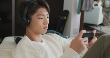 Kulaklık takan Asyalı çocuk akıllı telefonuyla oyun oynuyor evdeki kanepede oturuyor. Genç yaşam tarzı ve yaşam konsepti
