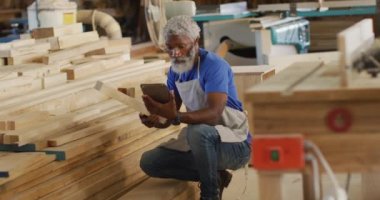 Dijital tabletli Afro-Amerikan erkek marangoz marangoz bir marangozhaneden ahşap tahta seçiyor. Marangozluk, işçilik ve el işi konsepti