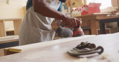 African american male carpenter wearing protective mask sanding a wooden plank with electric sander. carpentry, craftsmanship and handwork concept