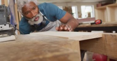 African american male carpenter blowing the dust over wooden plank at a carpentry shop. carpentry, craftsmanship and handwork concept